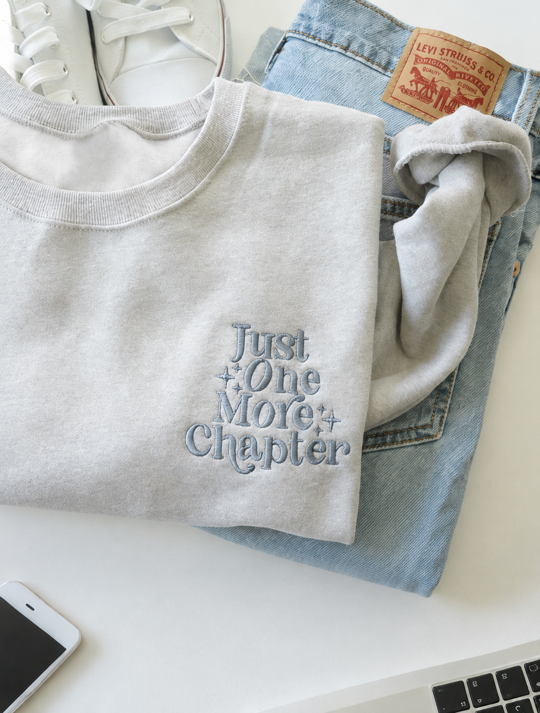 A light gray crewneck sweatshirt with the embroidered text 'Just One More Chapter' in blue-gray, styled in a flat-lay arrangement over light-wash blue jeans with a visible Levi's patch, alongside white sneakers and a smartphone on a white surface.