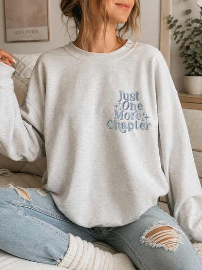 A woman wearing a light heather gray sweatshirt with the text 'Just One More Chapter' in blue embroidered lettering on the chest, paired with distressed light blue jeans.