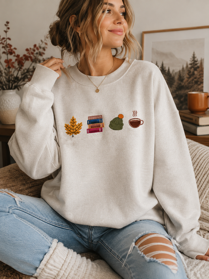 A woman sitting on a couch wearing a cream-colored crewneck sweatshirt with a small autumn-themed graphic on the chest, paired with distressed blue jeans.