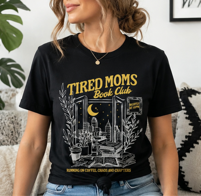Black t-shirt with 'Tired Moms Book Club' design worn by a woman, displayed with coffee and jeans.