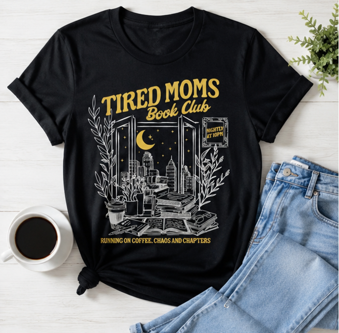 Black t-shirt with 'Tired Moms Book Club' design worn by a woman, displayed with coffee and jeans.